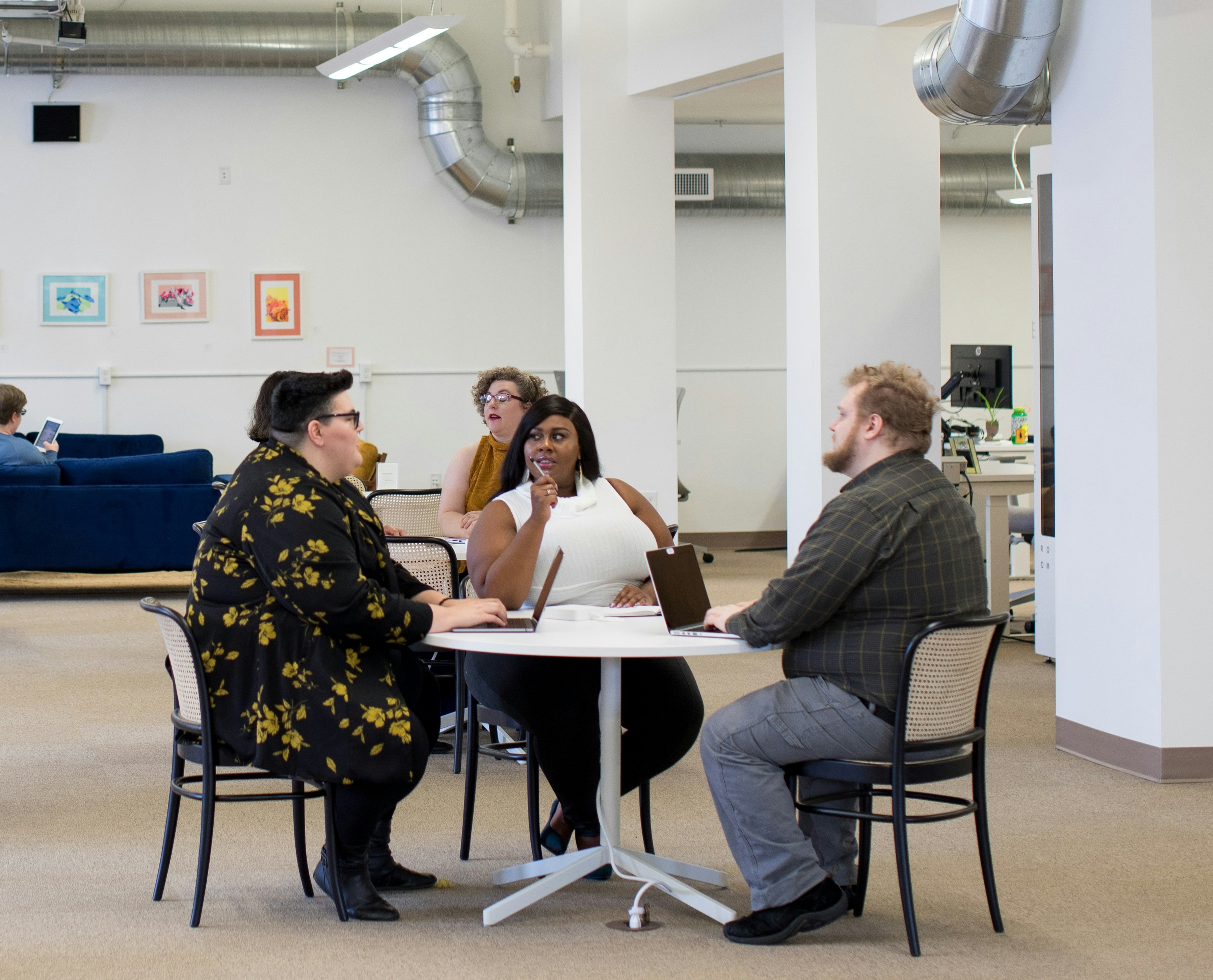plus size people sitting on chairs which look like kitchen chairs more than office chairs. Two people are white, one person is black and they are sitting around a table with laptops having a discussion in what could be the kitchen of a workplace