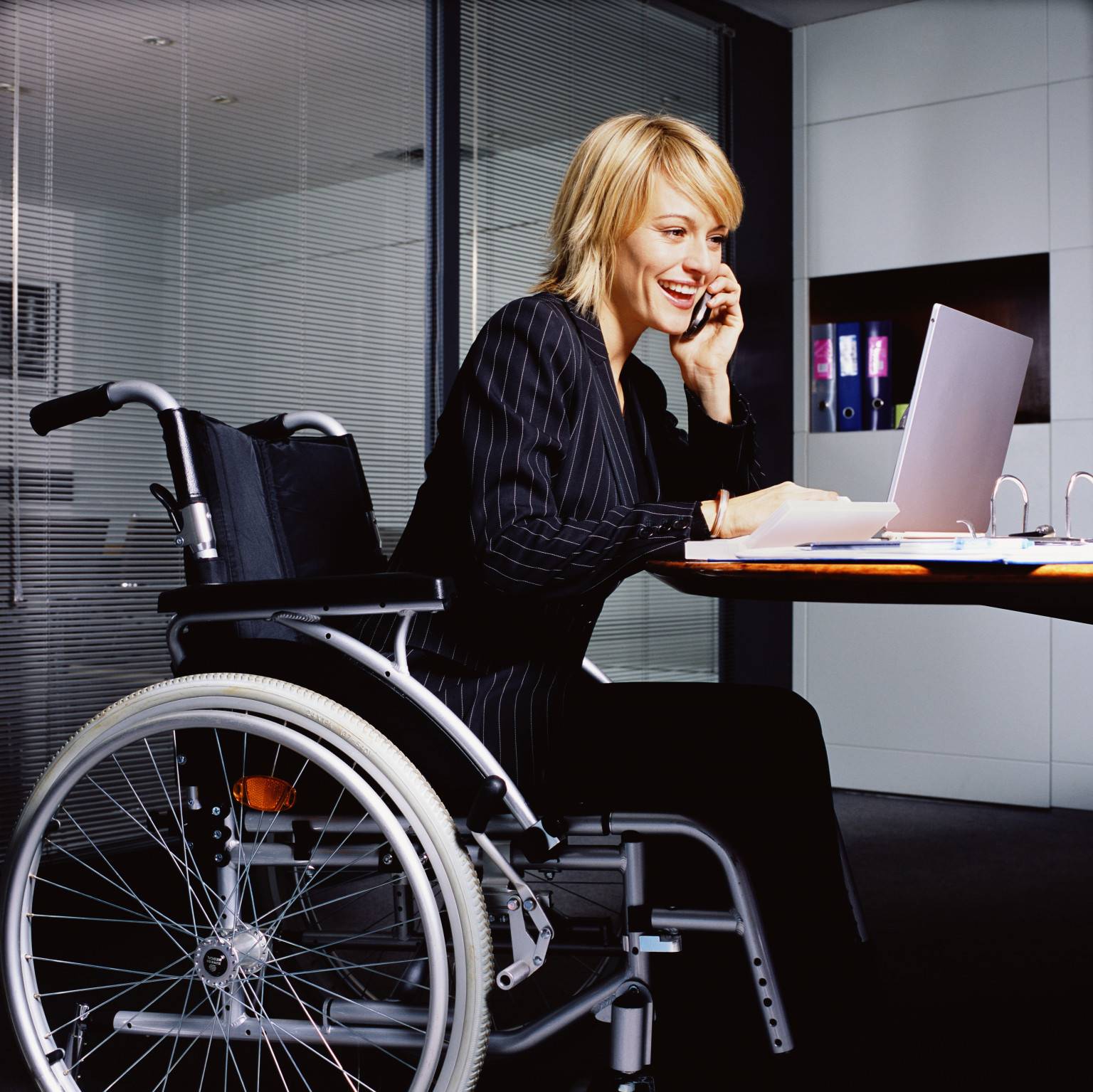 a young woman in a suit at her desk on the phone facing her computer at the office, sitting in a heavy wheelchair