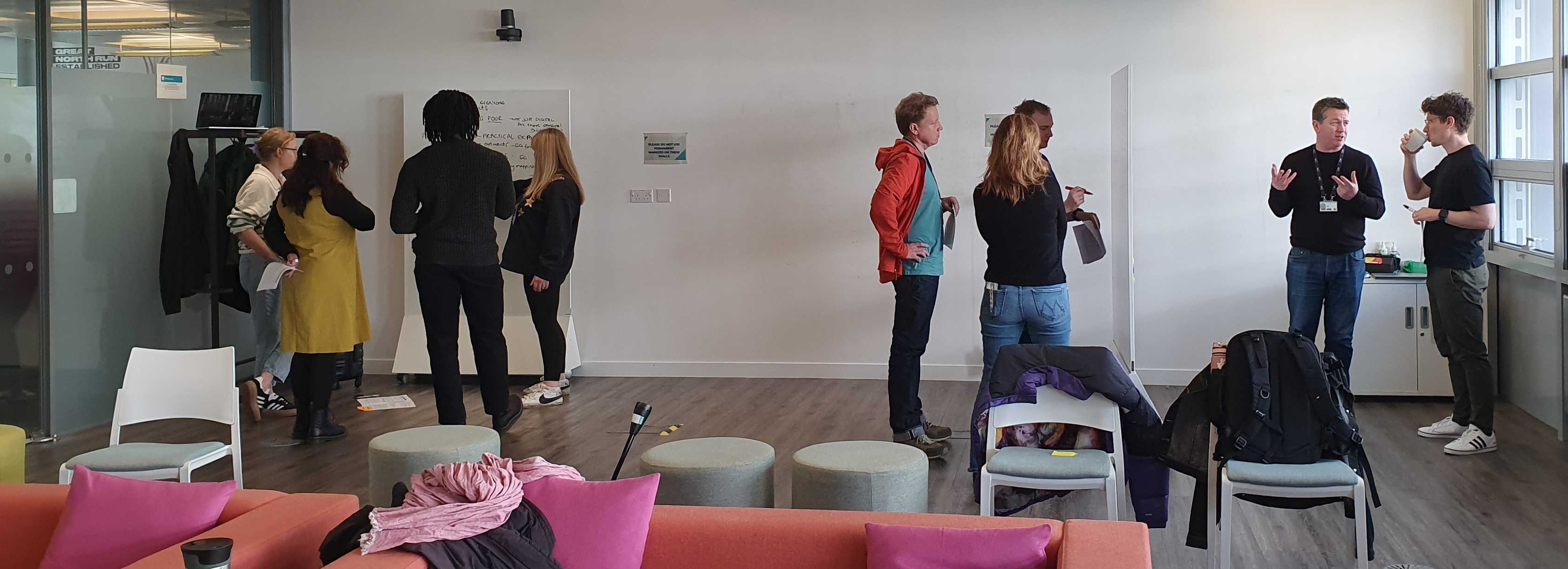 9 members of the service design community standing, working at whiteboards in an office room, with colourful sofa and cushions on the forefront